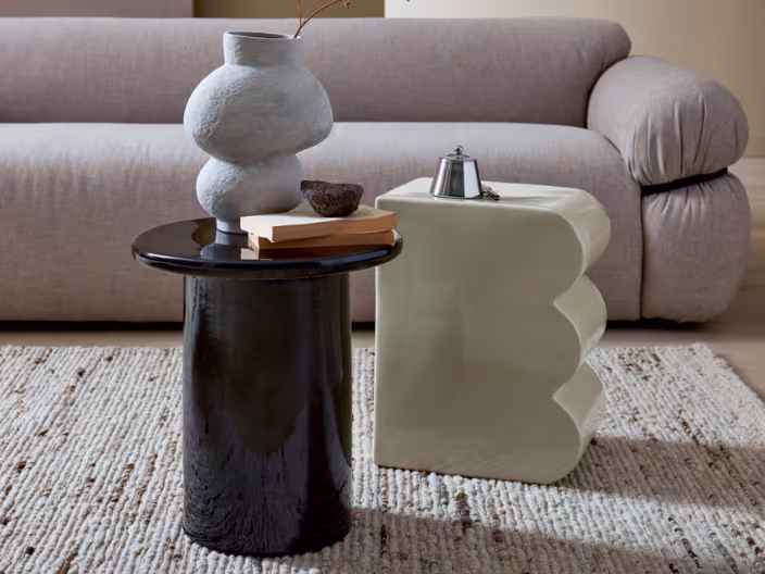 Moderne woonkamer met beige bank, zwarte en witte bijzettafels, een witte vaas en koffiepot op een natuurlijk tapijt.