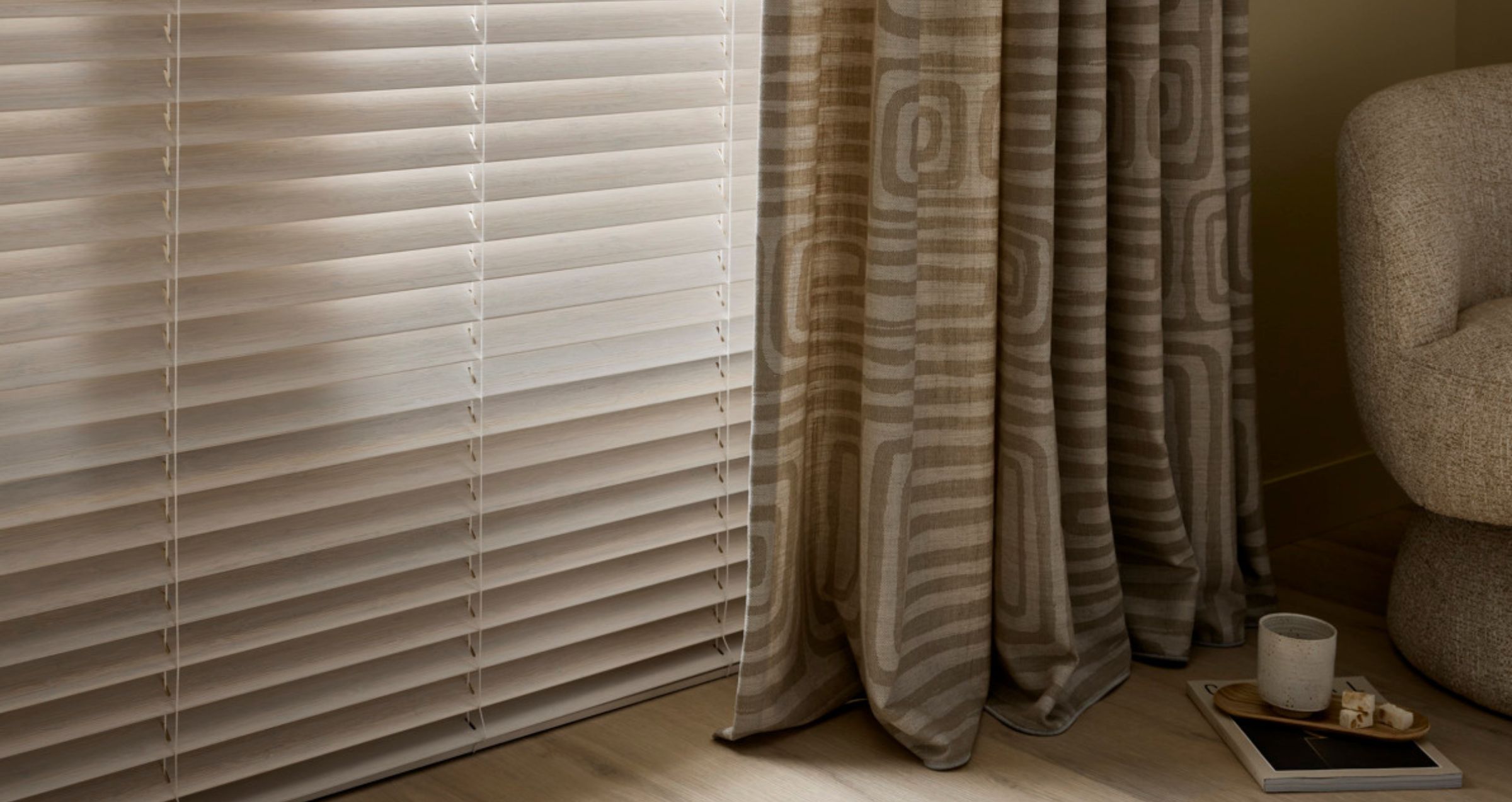 Witte jaloezieën naast beige gordijnen met geometrisch patroon in een woonkamer met een bank en klein dienblad.