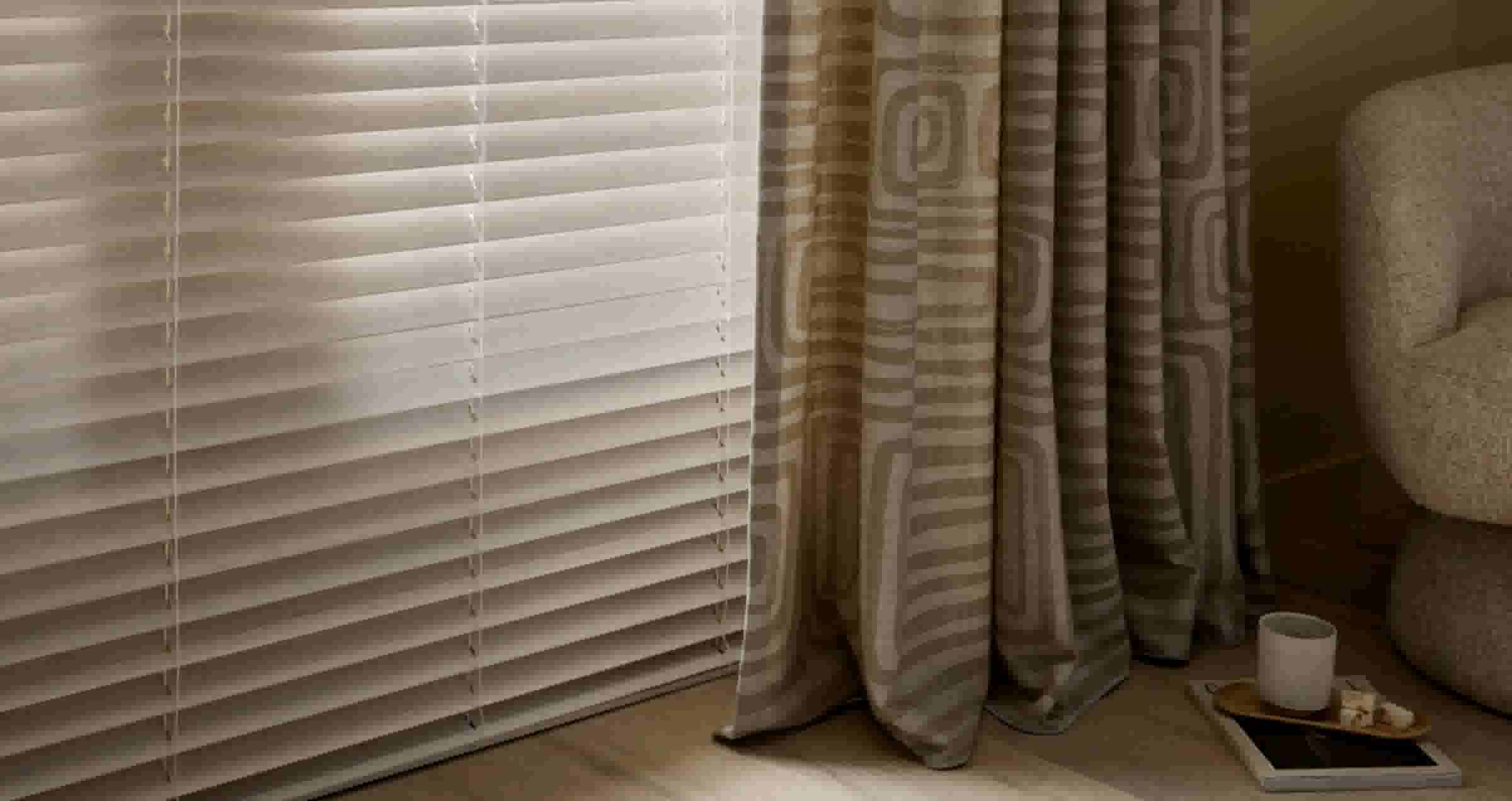 Witte jaloezieën naast beige gordijnen met geometrisch patroon in een woonkamer, met een kopje op een dienblad.