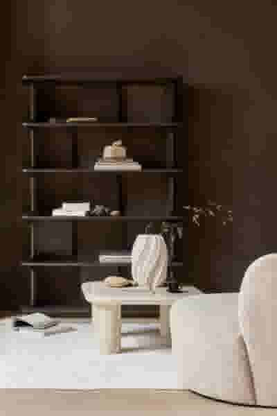 Minimalistisch interieur met donkere boekenplank, lichte salontafel met vaas en een crèmekleurige fauteuil op een wit tapijt.