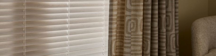 Witte horizontale jaloezieën naast beige gordijnen met geometrisch patroon in een rustige woonkamer.