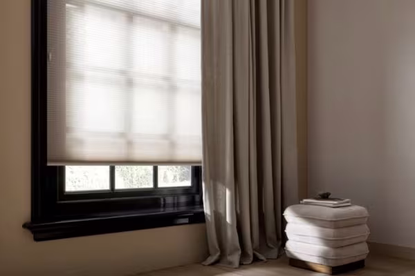 Raam met beige plisségordijn en linnen overgordijn. Naast het raam staat een witte poef met boeken, in een rustige kamer.