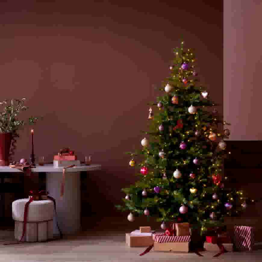 Versierde kerstboom met kleurrijke ballen en lichtjes, omringd door cadeaus, naast een tafel met kaars en plant in gezellige kamer.
