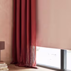 Roestbruine gordijnen naast een rolgordijn bij een raam, met een roze muur en een lamp met boeken op de achtergrond.