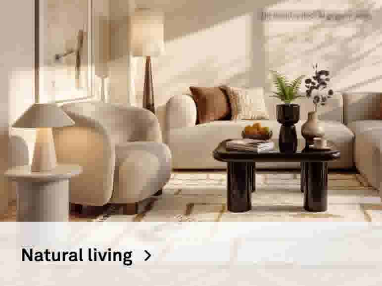 Natural living. Sfeerbeeld van een beige fauteuil en bank met sierkussens, een donkere salontafel met vazen en schaal fruit, tafellamp en vloerlamp, op een crème vloerkleed.