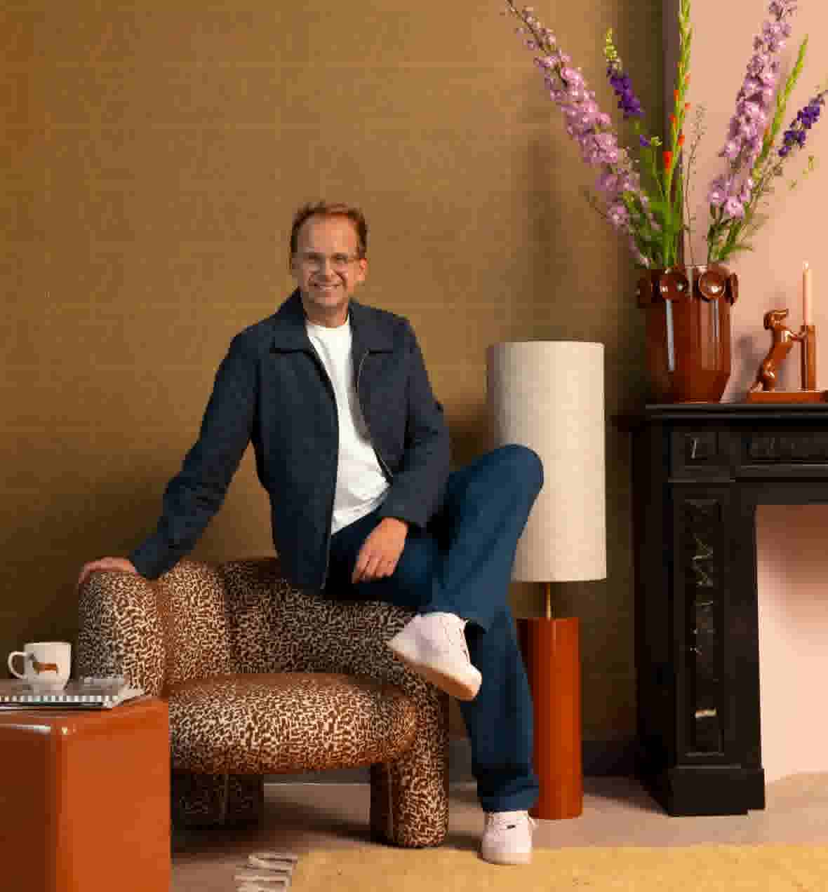 Sfeerbeeld van Theo-Bert Pot, zittend op een bruin met wit gepatroonde poef, met een vaas bloemen, kandelaar en teckelbeeldje op een zwarte schouw.