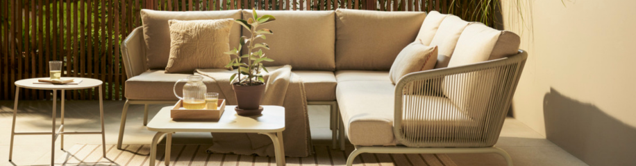 Gezellige buitenhoek met beige loungebank, kussens, salontafel met plant en dienblad, en bijzettafel in zonnige tuin.