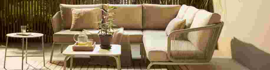 Gezellige buitenhoek met beige loungebank, kussens, salontafel met plant en dienblad, en bijzettafel in zonnige tuin.