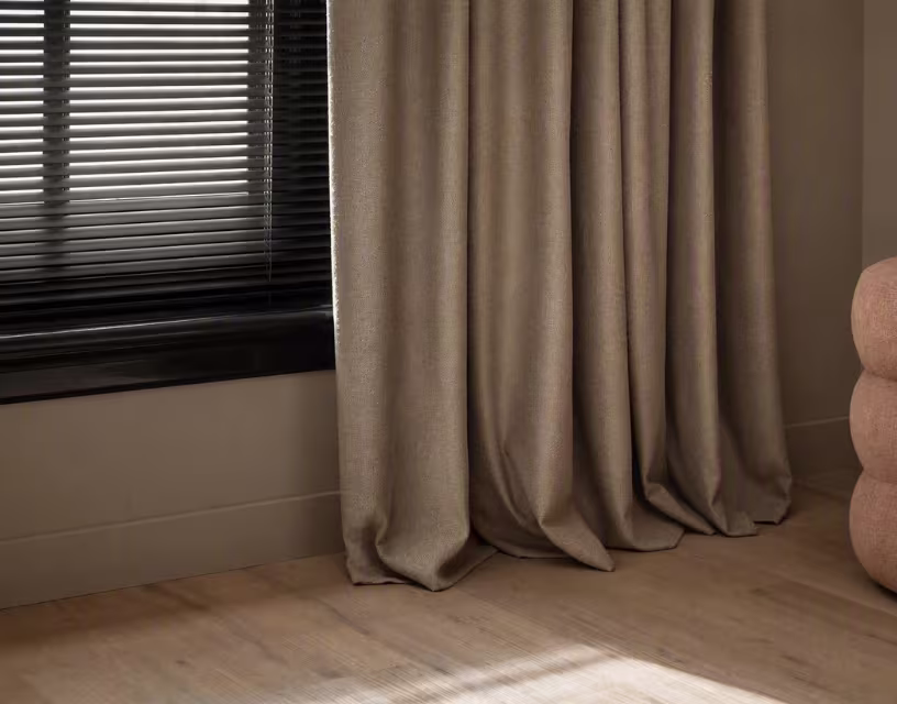 Taupe gordijnen hangen voor een raam met zwarte jaloezieën in een moderne kamer met houten vloer en beige muur.