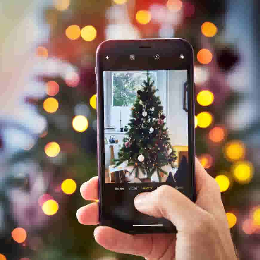 Iemand maakt een foto van een versierde kerstboom met een smartphone, tegen een achtergrond van feestelijke lichtjes.