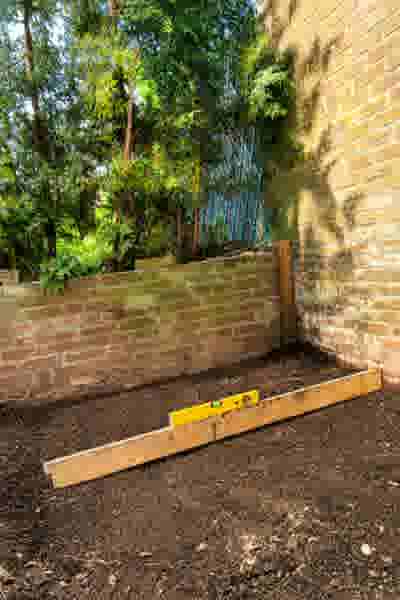 Een houten plank met een geel waterpas ligt op omgewoelde aarde in een tuin, omgeven door bakstenen muren en groen.