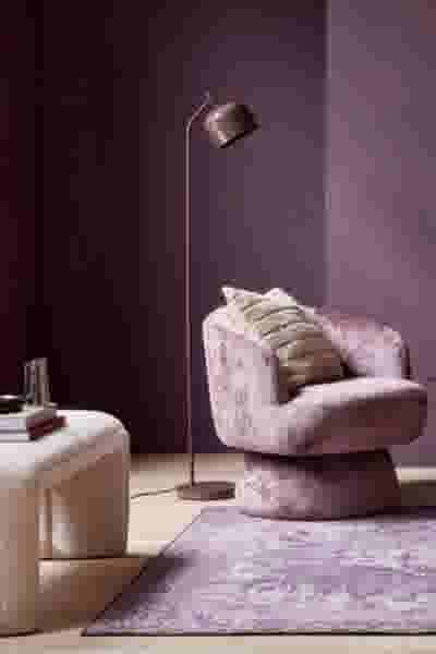 Moderne woonkamer met paarse muren, roze fluwelen fauteuil, bronzen staande lamp en crèmekleurige poef op een paars tapijt.