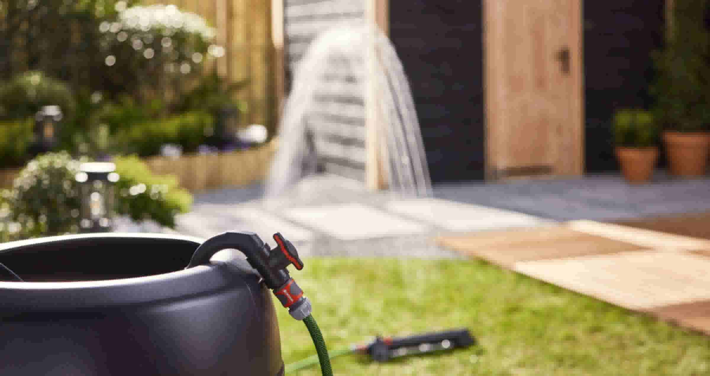 Zwarte regenton met groene tuinslang aangesloten op kraan. Op de achtergrond sproeit water boogvormig in de tuin.