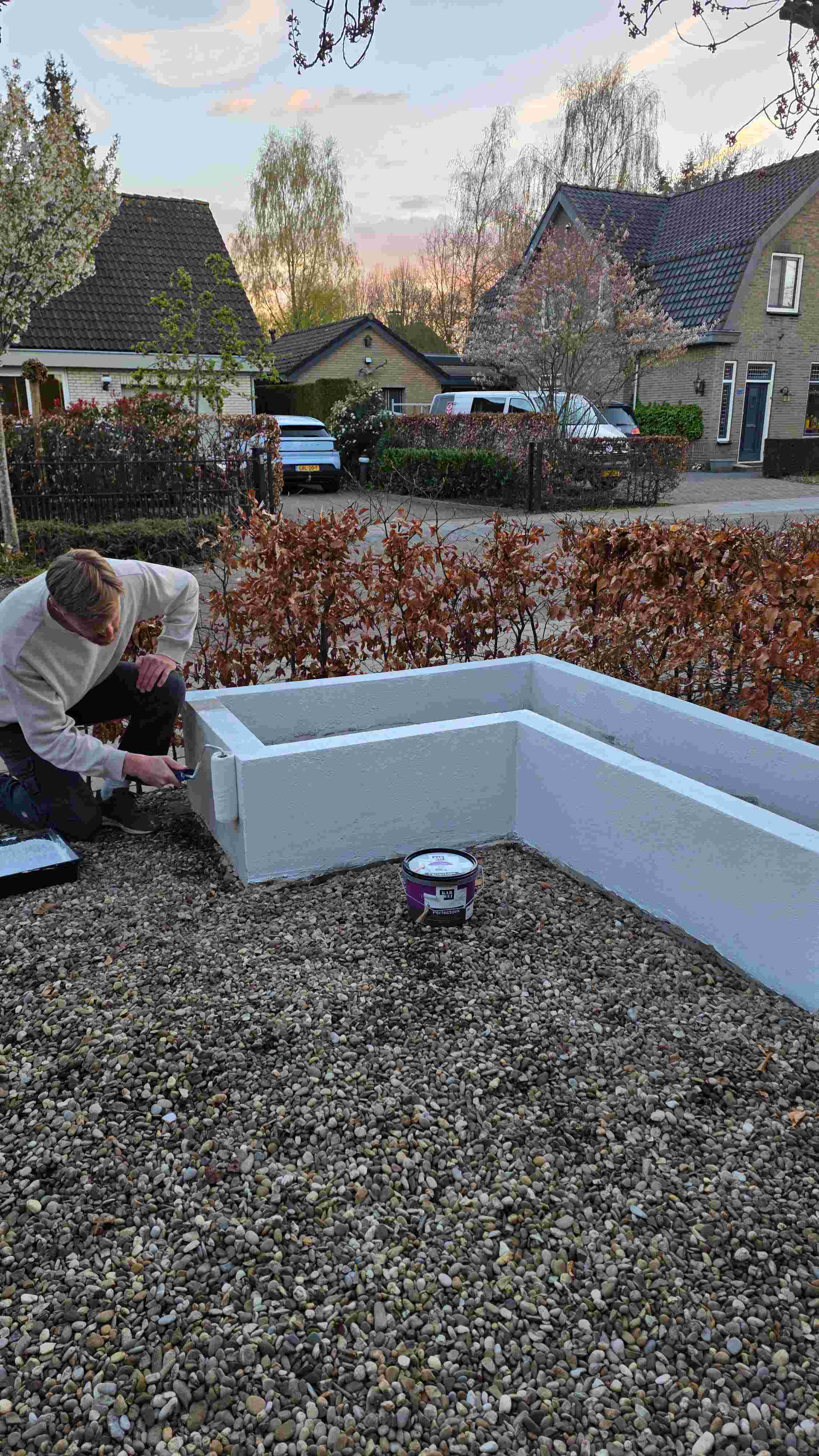 Persoon schildert een witte muurbak in een tuin met kiezelstenen, met huizen en bloeiende bomen op de achtergrond bij zonsondergang.