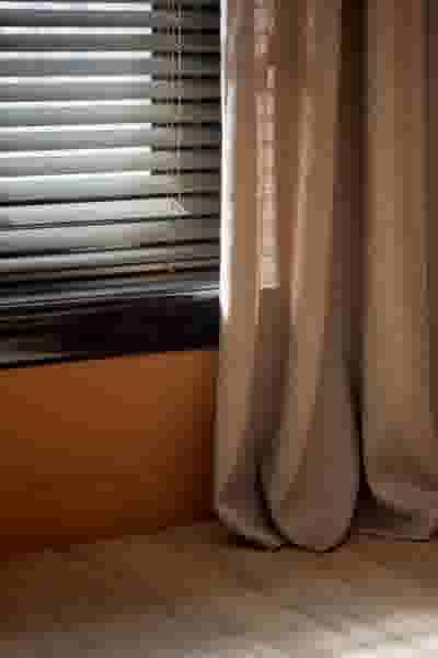 Raam met aluminium jaloezieën en beige gordijnen die tot op de houten vloer hangen, naast een oranje muur.