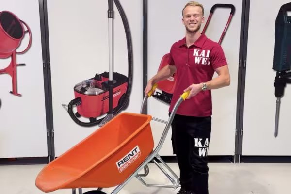 Karwei medewerker met een oranje RentPartner kruiwagen in een showroom met bouwgereedschap op de achtergrond.
