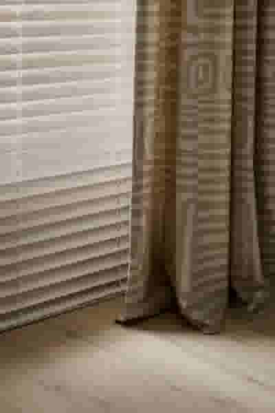 Witte jaloezieën naast beige gordijnen met geometrisch patroon op een lichte houten vloer in een rustig interieur.