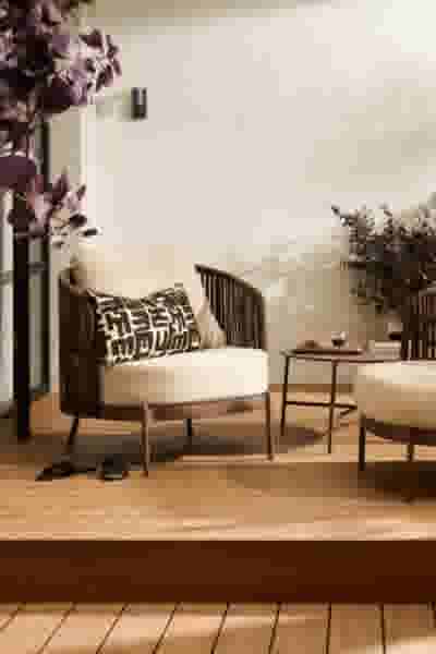 Gezellige zithoek in de tuin met crèmekleurige fauteuil, bijzettafel en poef op houten vloer, versierd met paarse planten.