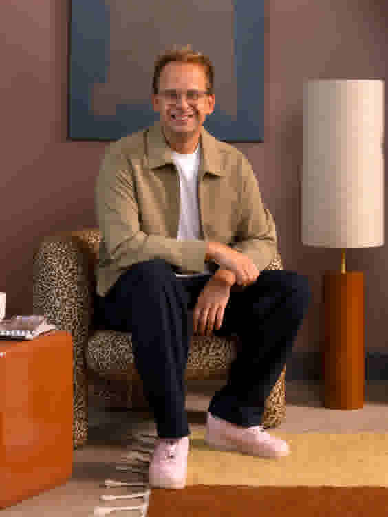 Sfeerbeeld van Theo-Bert Pot, glimlachend gezeten op een bruin met wit gepatroonde fauteuil, met een vloerlamp en oranje bijzettafel, voor een blauw-bruin schilderij, op een geel-oranje vloerkleed.