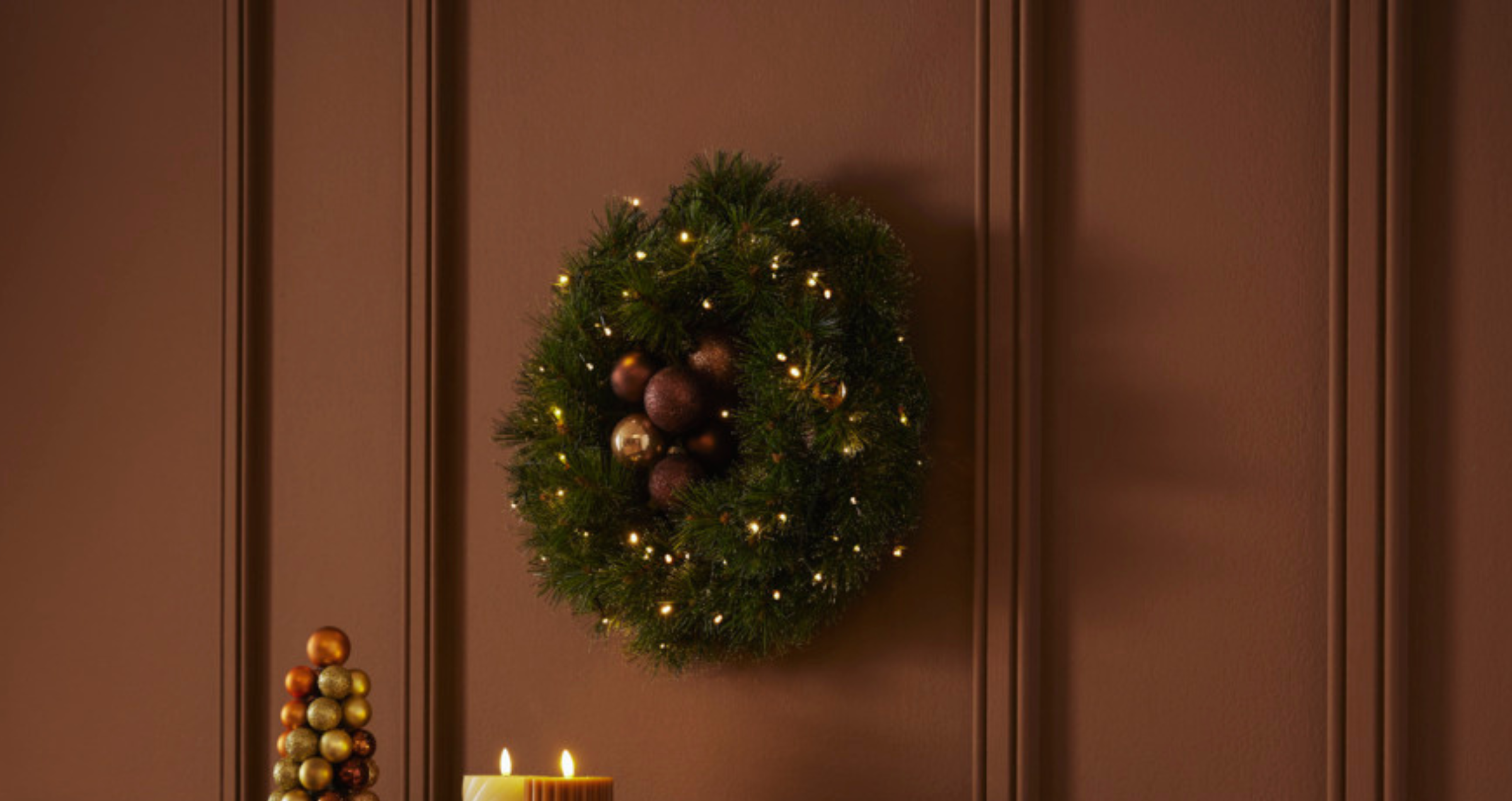 Feestelijke kerstkrans met lichtjes en ballen hangt aan bruine muur, met decoratieve kerstboom en kaarsen eronder.