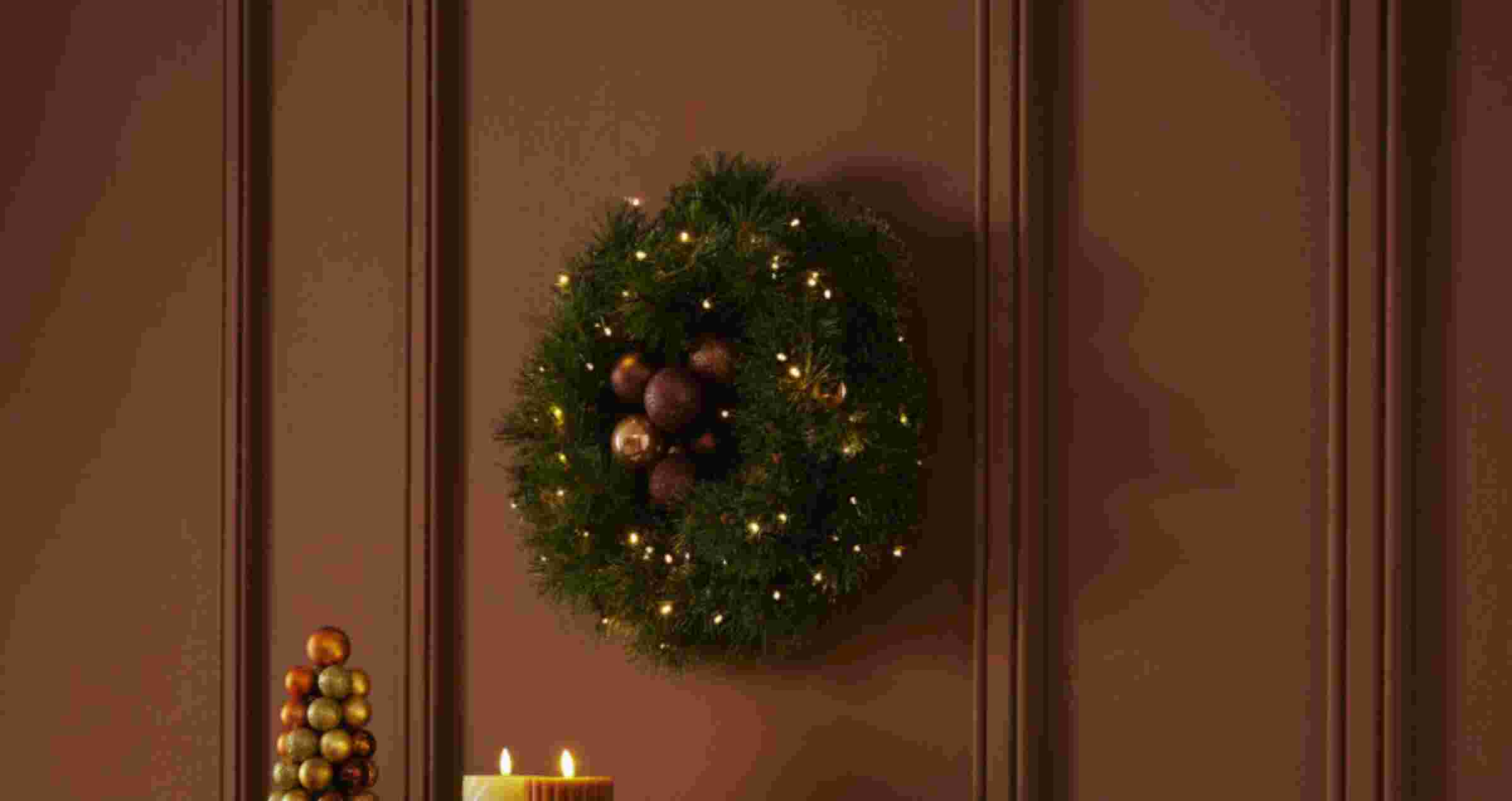 Feestelijke kerstkrans met lichtjes en ballen hangt aan bruine muur, met decoratieve kerstboom en kaarsen eronder.