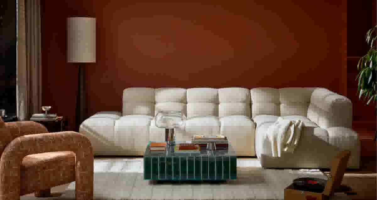 Stijlvolle woonkamer met cremekleurige modulaire bank, groene salontafel met boeken, oranje fauteuil en terracotta muur.