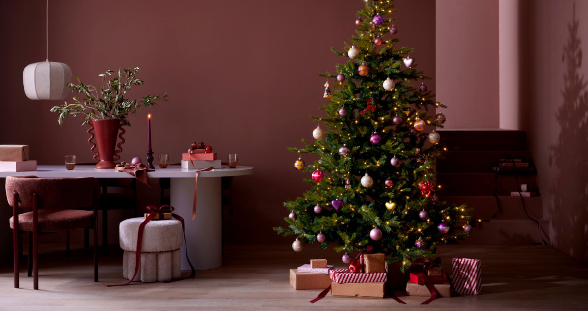 Versierde kerstboom met kleurrijke ballen en lichtjes naast een eettafel in een kamer met bordeauxrode muren en cadeaus eronder.