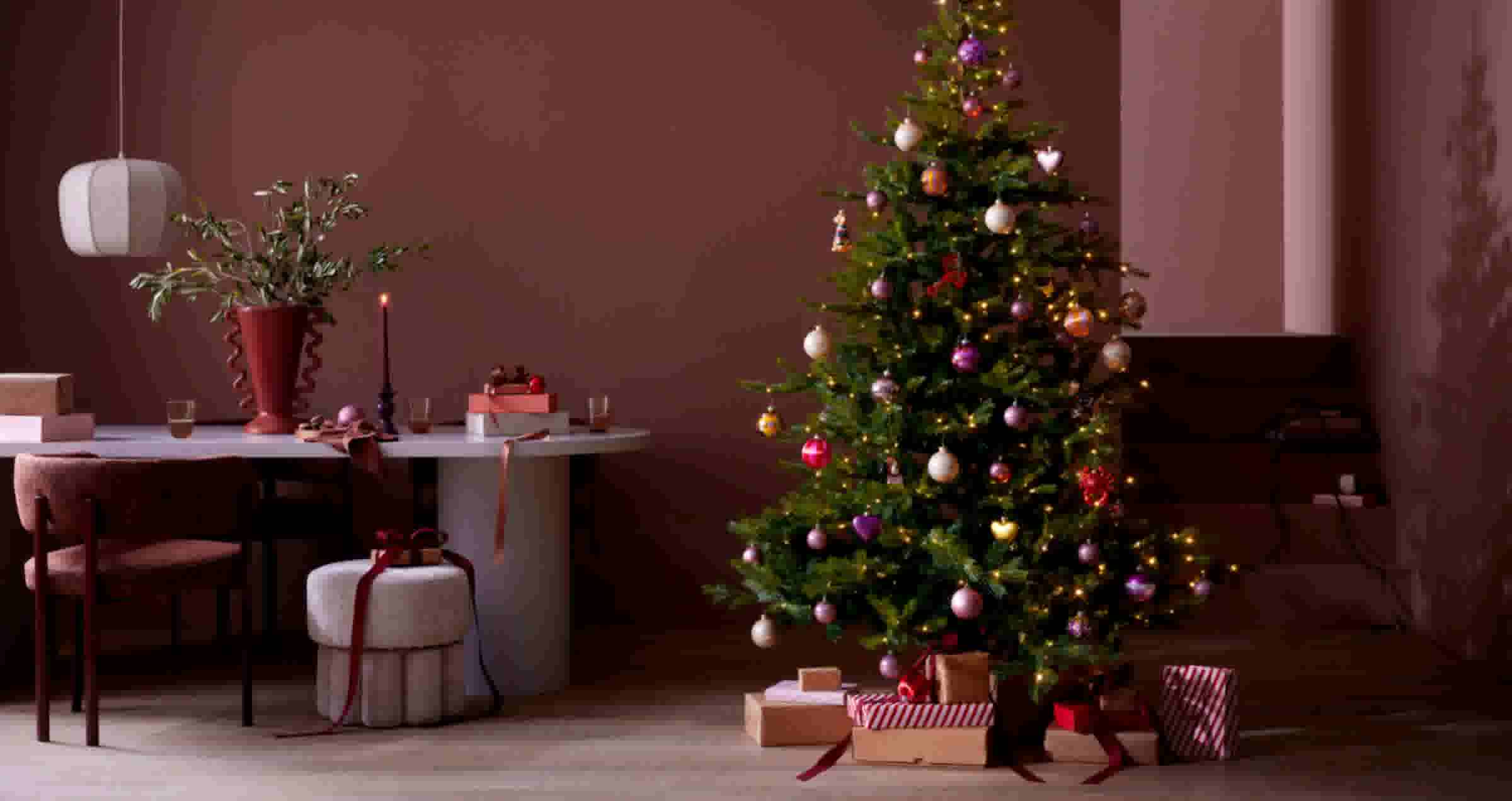 Versierde kerstboom met kleurrijke ballen en lichtjes naast een eettafel in een stijlvolle woonkamer met bordeauxrode muren.