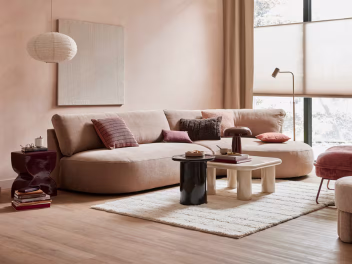 Warme woonkamer met beige hoekbank, kleurrijke kussens, ronde salontafels op wit tapijt en roze muren.