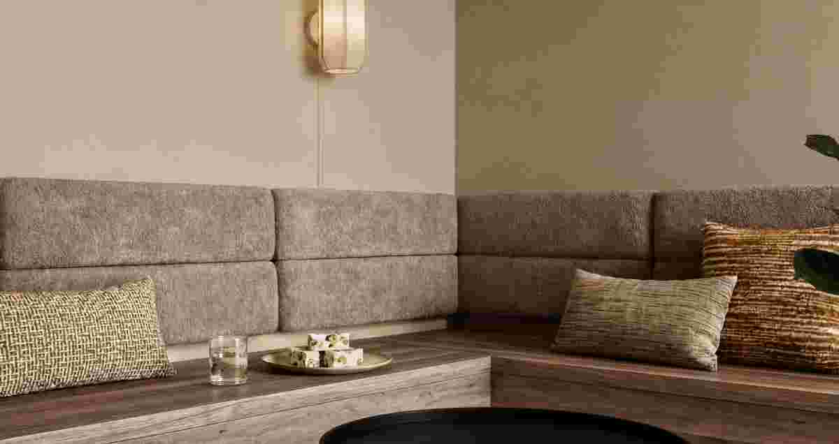 Grijze hoekbank met sierkussens, houten tafel met glas water en hapjes op gouden schaaltje, wandlamp aan beige muur.