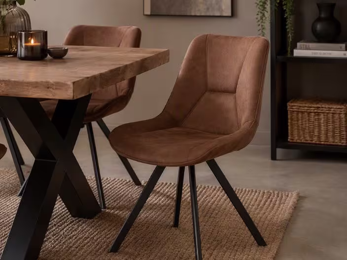 Bruine leren eetkamerstoel met zwarte poten naast een houten tafel met X-poten, op een juten vloerkleed in een warme eetkamer.