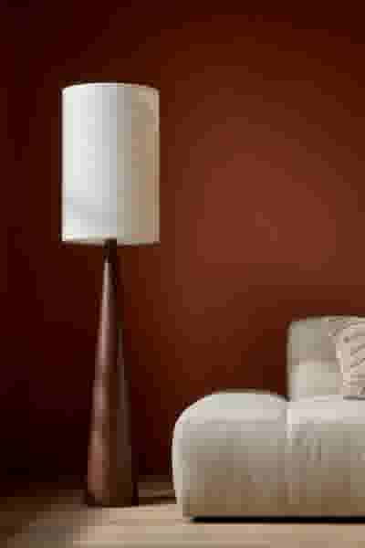 Moderne staande lamp met houten voet en witte kap naast een lichte bank tegen een warme terracotta muur.