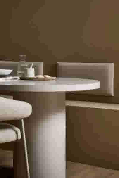 Minimalistisch eethoek met ronde witte tafel, beige zitbank en stoel tegen een bruine muur met wandkussens in een cremekleur.