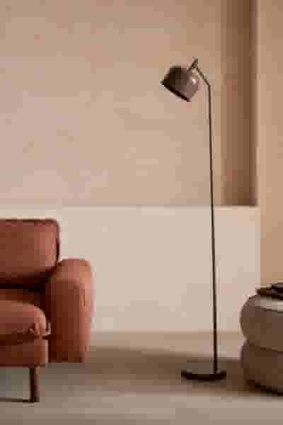 Slanke vloerlamp met bruine kap op zwart frame, naast een terracotta bank en ronde poef in een warme, beige ruimte.