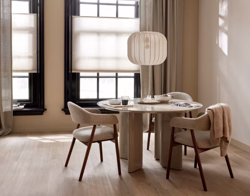 Moderne eetkamer met ronde tafel, beige stoelen en hanglamp bij groot raam met zwarte kozijnen en neutrale tinten.