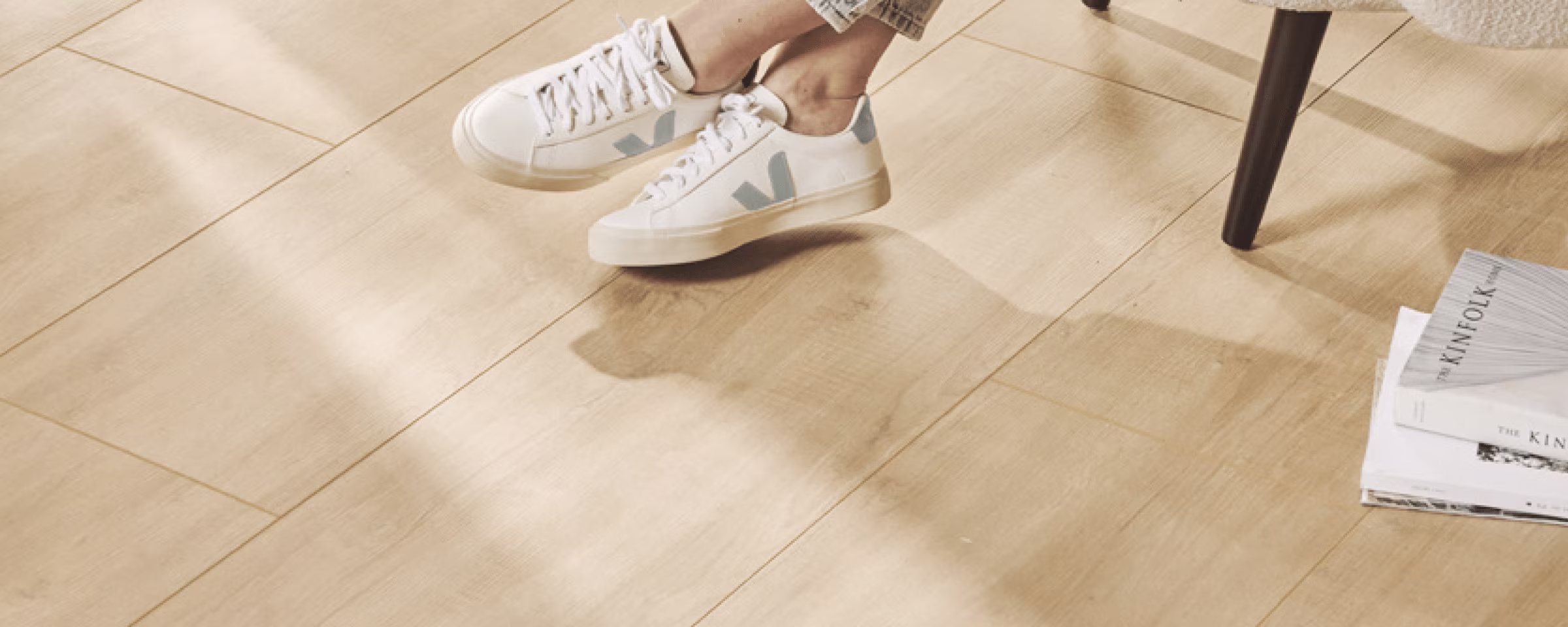 Persoon in spijkerbroek en grijze trui draagt witte sneakers met groene accenten op lichte houten vloer bij stoel.