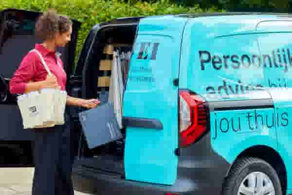 Persoon in roze shirt met tas haalt documenten uit een turquoise bedrijfsbusje met "Persoonlijk advies bij jou thuis" erop.