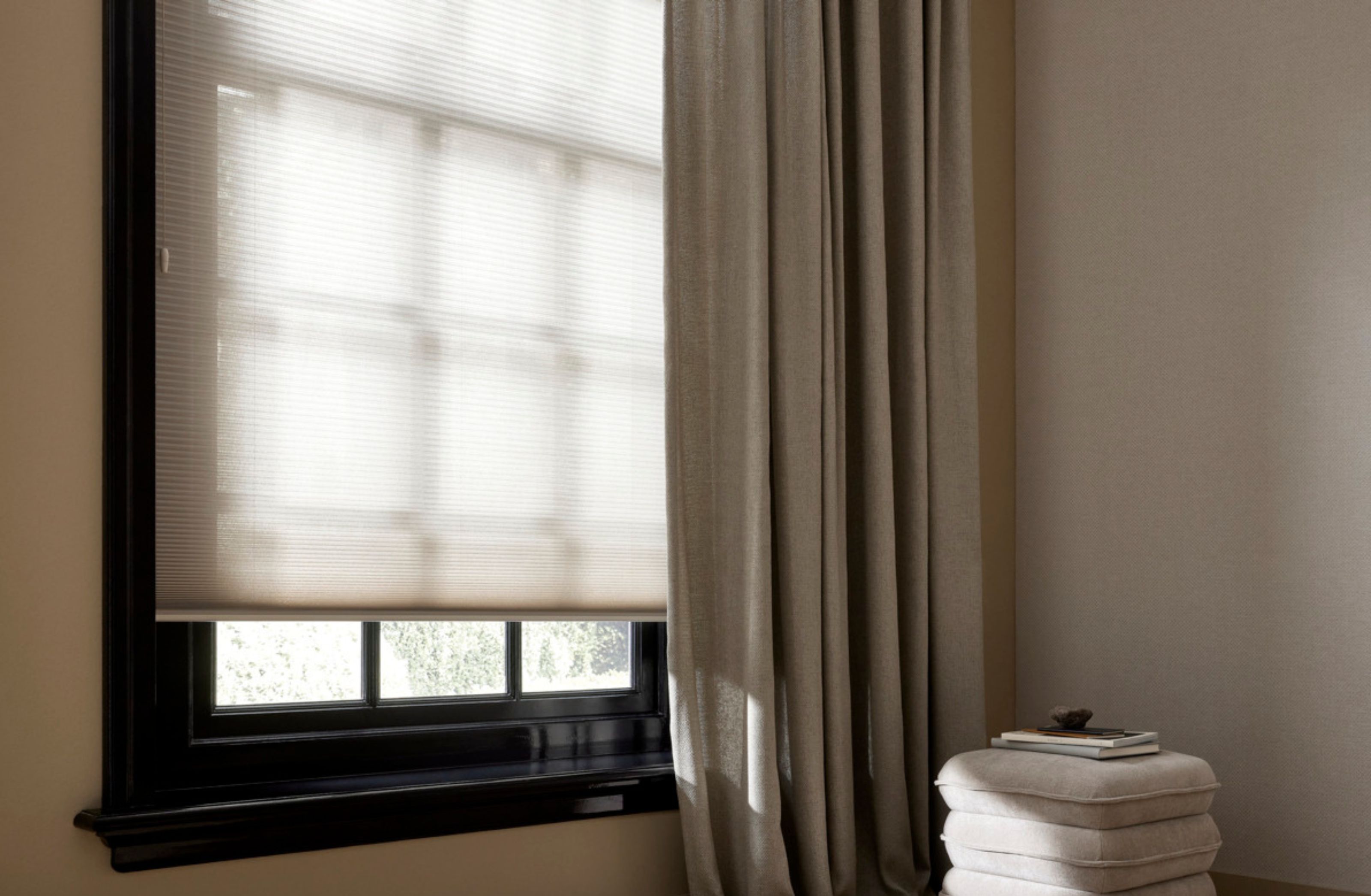 Moderne woonkamer met beige gordijnen en rolgordijn bij raam, witte bijzettafel met kaars en boek op houten vloer.