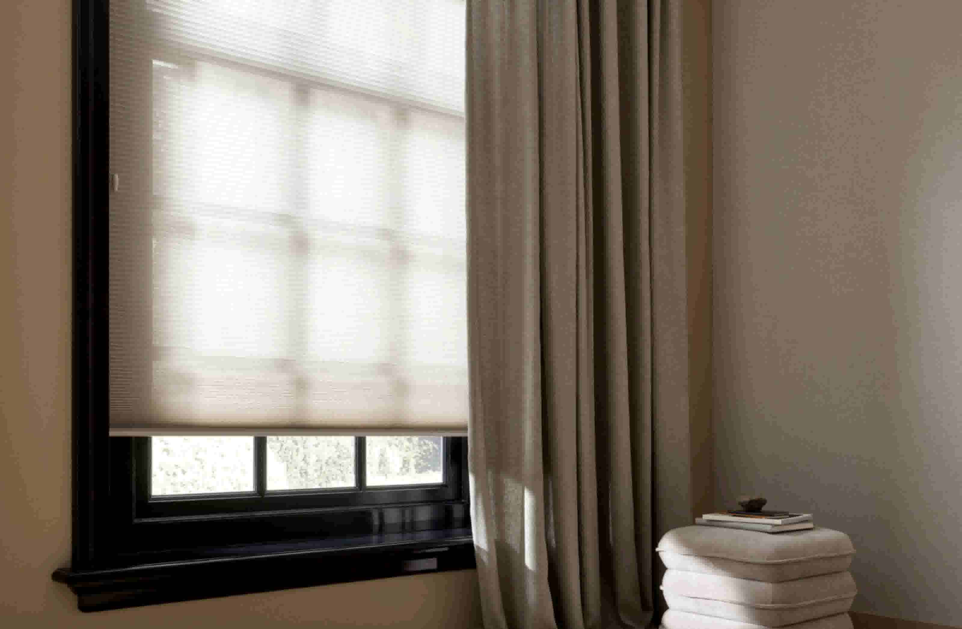 Moderne woonkamer met beige gordijnen en rolgordijn bij raam, witte bijzettafel met kaars en boek op houten vloer.