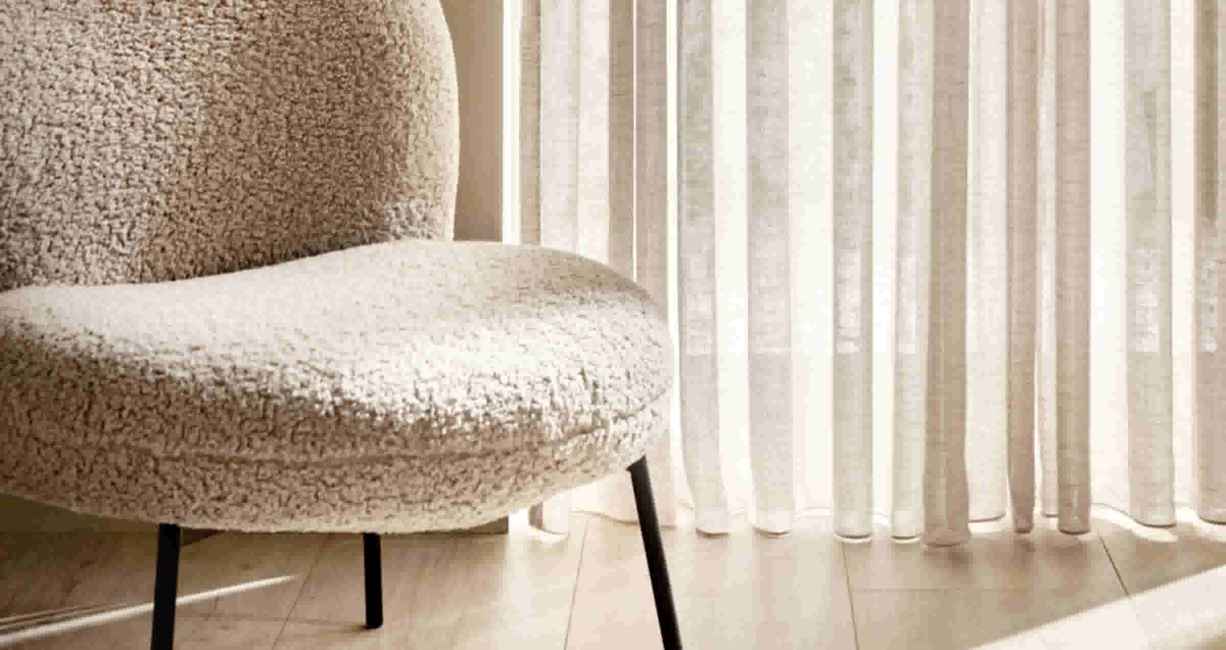 Beige teddy-stof fauteuil met zwarte poten naast lichte linnen gordijnen in een minimalistisch interieur.