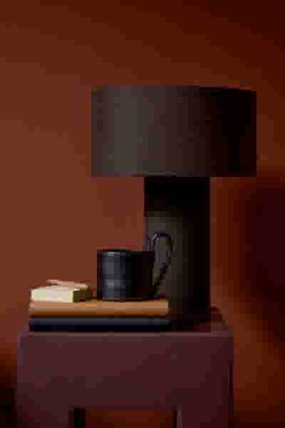 Moderne donkerbruine tafellamp met ronde kap op een bruine tafel, naast een zwarte mok en gestapelde boeken in warme tinten.