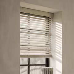 Witte houten jaloezie voor een raam, zonlicht werpt schaduwstrepen op de muur in een lichte kamer.