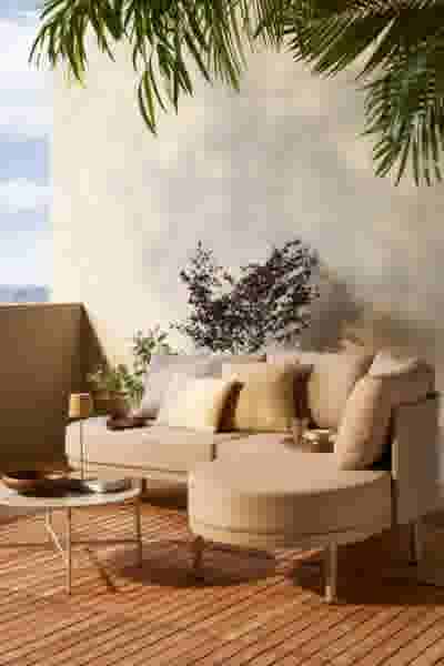 Beige loungebank met kussens op houten terras, omringd door palmbladeren en een kleine plant in een tropische sfeer.