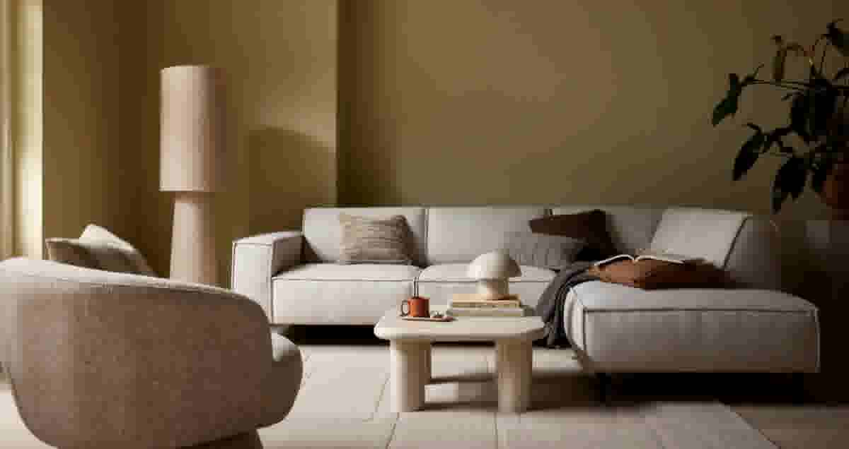 Moderne woonkamer met lichtgrijze hoekbank, beige salontafel, vloerlamp en plant tegen olijfgroene muur.
