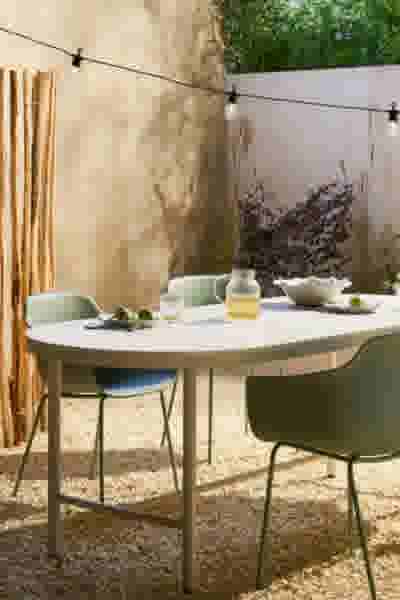Buiteneethoek met witte ovale tafel en groene stoelen op grind, omgeven door planten en slingerverlichting.