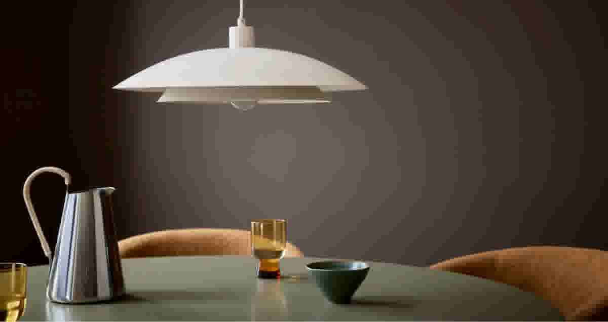 Witte hanglamp boven een groene tafel met een zilveren kan, amber glas en een blauw kommetje. Bruine stoelen op de achtergrond.