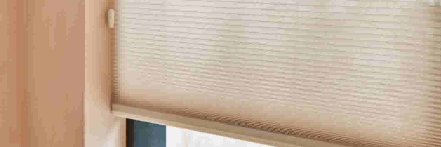 Beige honingraat rolgordijn gedeeltelijk neergelaten voor een raam, met een subtiel gestreept textuurpatroon.