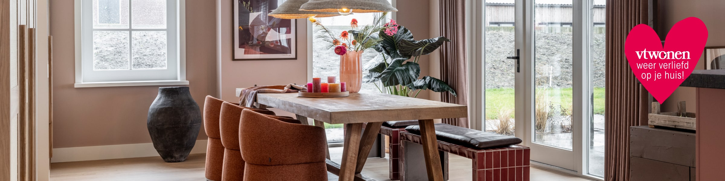 Gezellige eetkamer met houten tafel, roestbruine stoelen en bankje, verse bloemen in vaas, kaarsen en grote ramen met uitzicht op tuin.