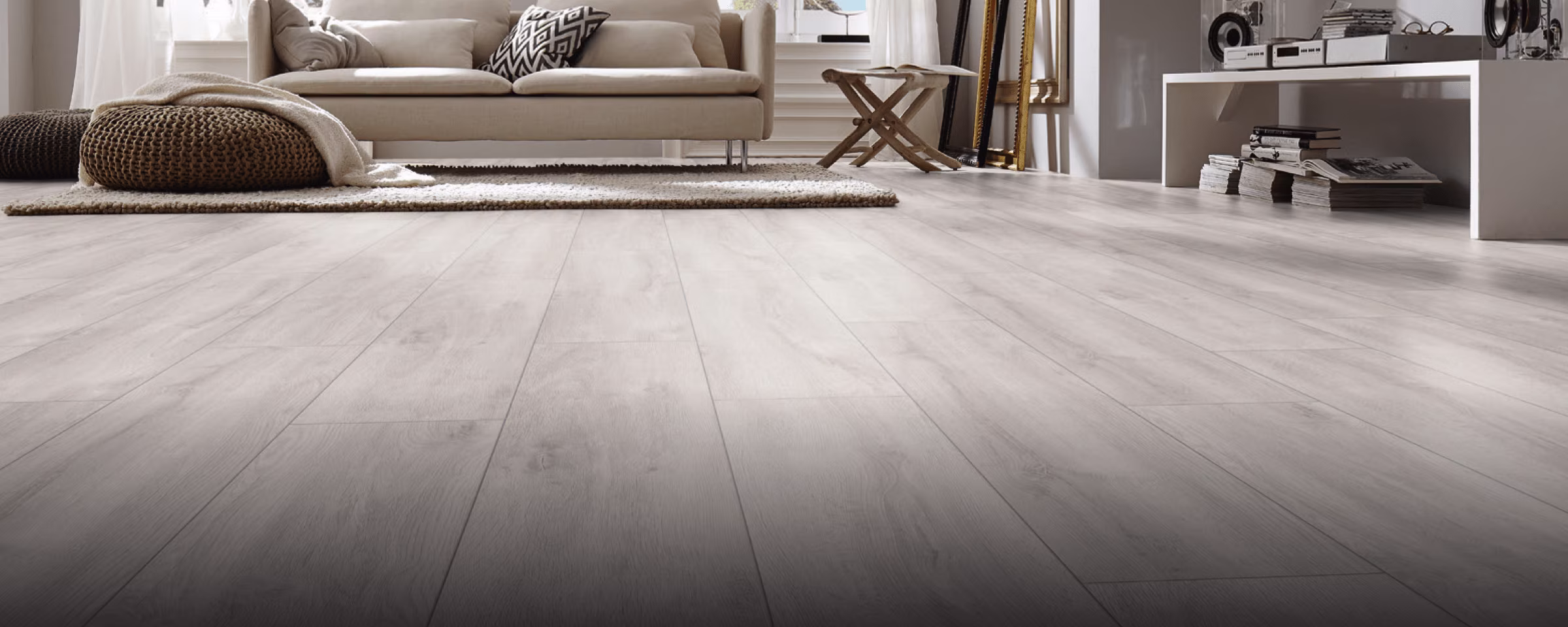 Moderne woonkamer met lichte laminaatvloer, beige bank, gevlochten poef, houten bijzettafel en witte boekenkast.