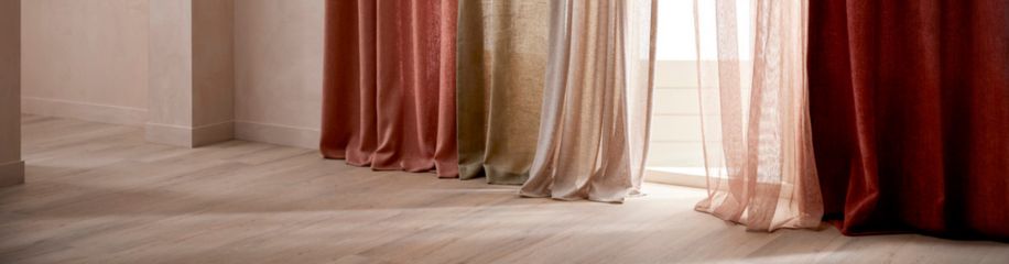 Elegante gordijnen in warme aardetinten van rood en beige hangen tot op de lichte houten vloer in een lichte kamer.